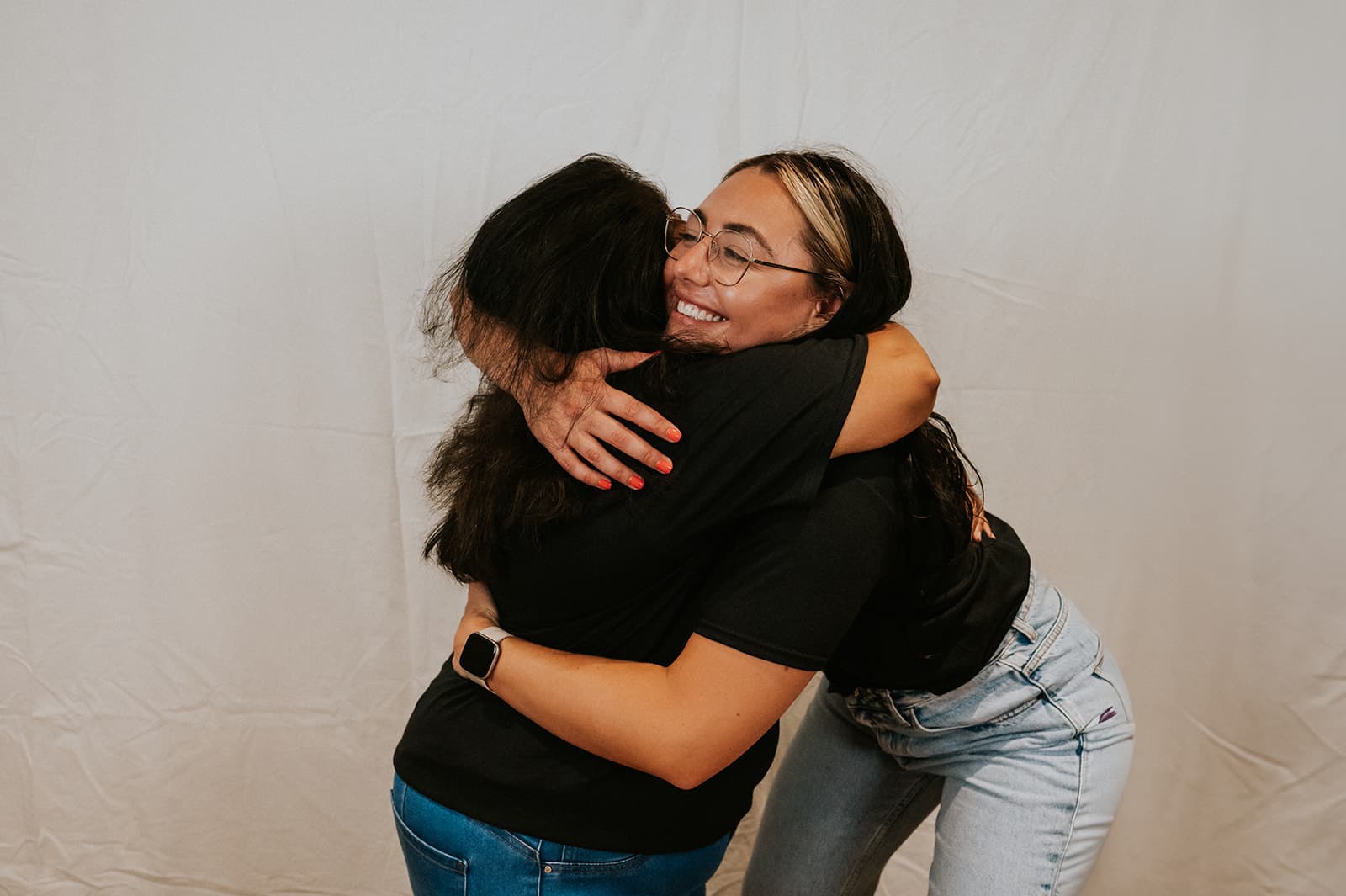 Nicole, wearing glasses and a black ‘The Women’s Wellbeing Club’ t-shirt, sits against a softly lit wall, playfully running her hands through her hair. A warm-glowing floor lamp stands to her left, casting an inviting ambiance.
