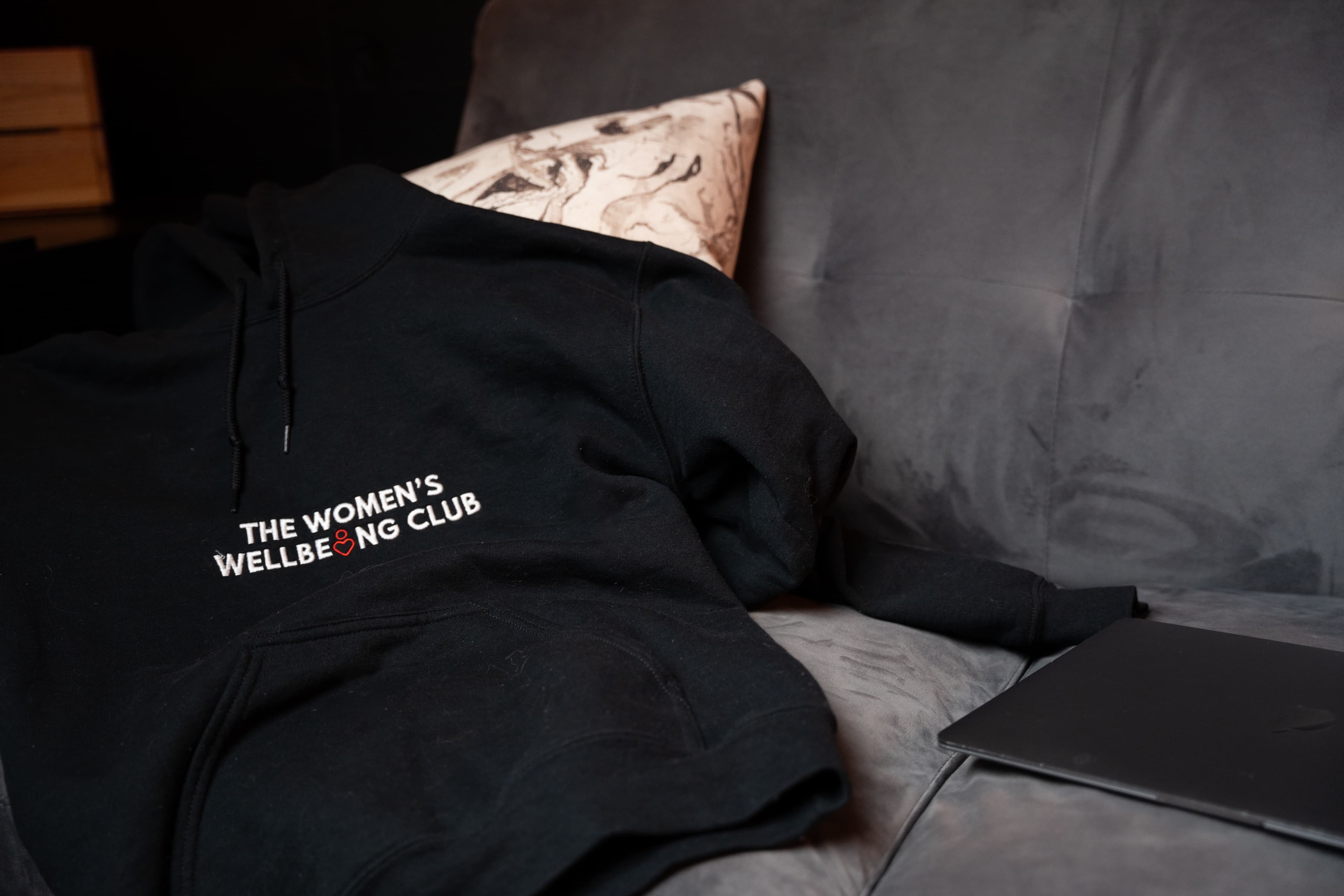 Nicole, wearing glasses and a black ‘The Women’s Wellbeing Club’ t-shirt, sits against a softly lit wall, playfully running her hands through her hair. A warm-glowing floor lamp stands to her left, casting an inviting ambiance.