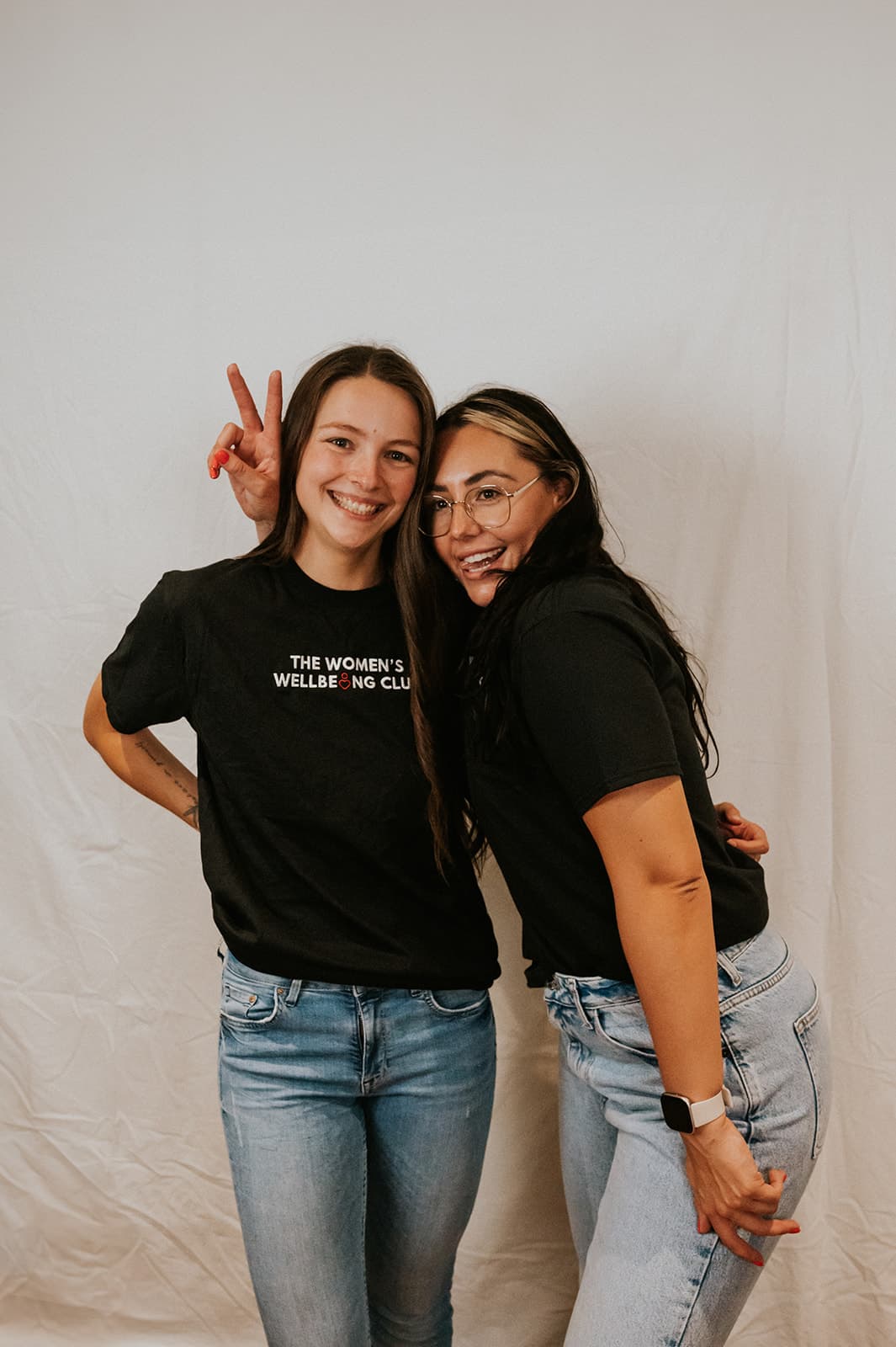 Nicole, wearing glasses and a black ‘The Women’s Wellbeing Club’ t-shirt, sits against a white wall with a warm light to the side. She smiles confidently while raising one leg toward the camera, showcasing the sole of her Vans sneaker in a playful, dynamic pose.