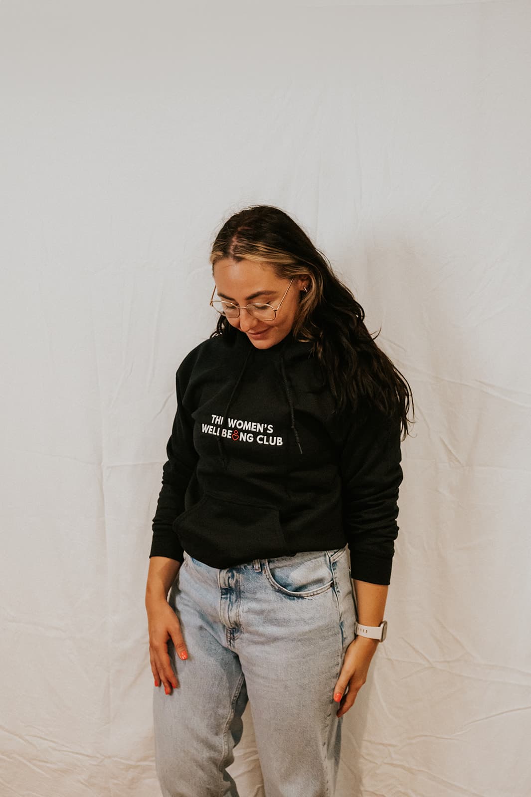 Nicole, wearing glasses and a black 'The Women's Wellbeing Club' t-shirt, sits on a dark grey couch working on a laptop.